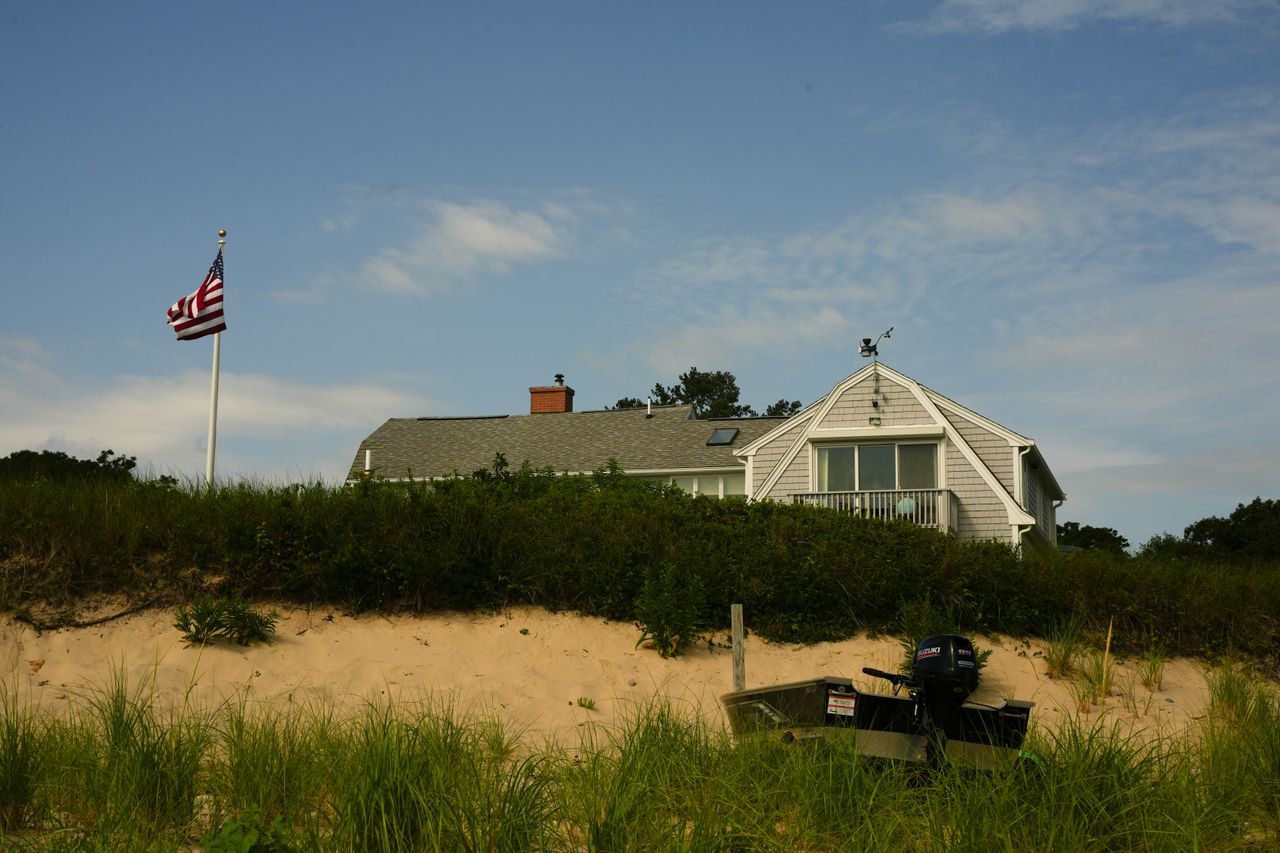 expertise-01 A picturesque beach house with an American flag, surrounded by dune grass on a sunny day.